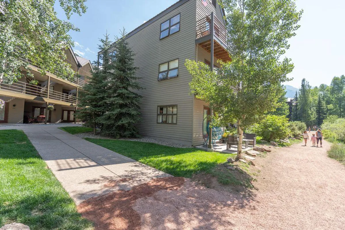 A modern condominium building framed by lush greenery and set along a walking path. The scene is calm and inviting, suggesting a safe and scenic spot for families to stroll, bike, or let children explore right outside the property. The path hints at immediate access to outdoor recreation and the river trail mentioned in the section.
