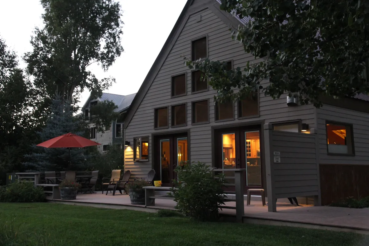 The image features a spacious house with large windows and a generous outdoor seating area under a vibrant red umbrella. The atmosphere is cozy and inviting, perfect for gatherings or enjoying meals outside. The surrounding area is serene, with hints of greenery and ample space for group activities.