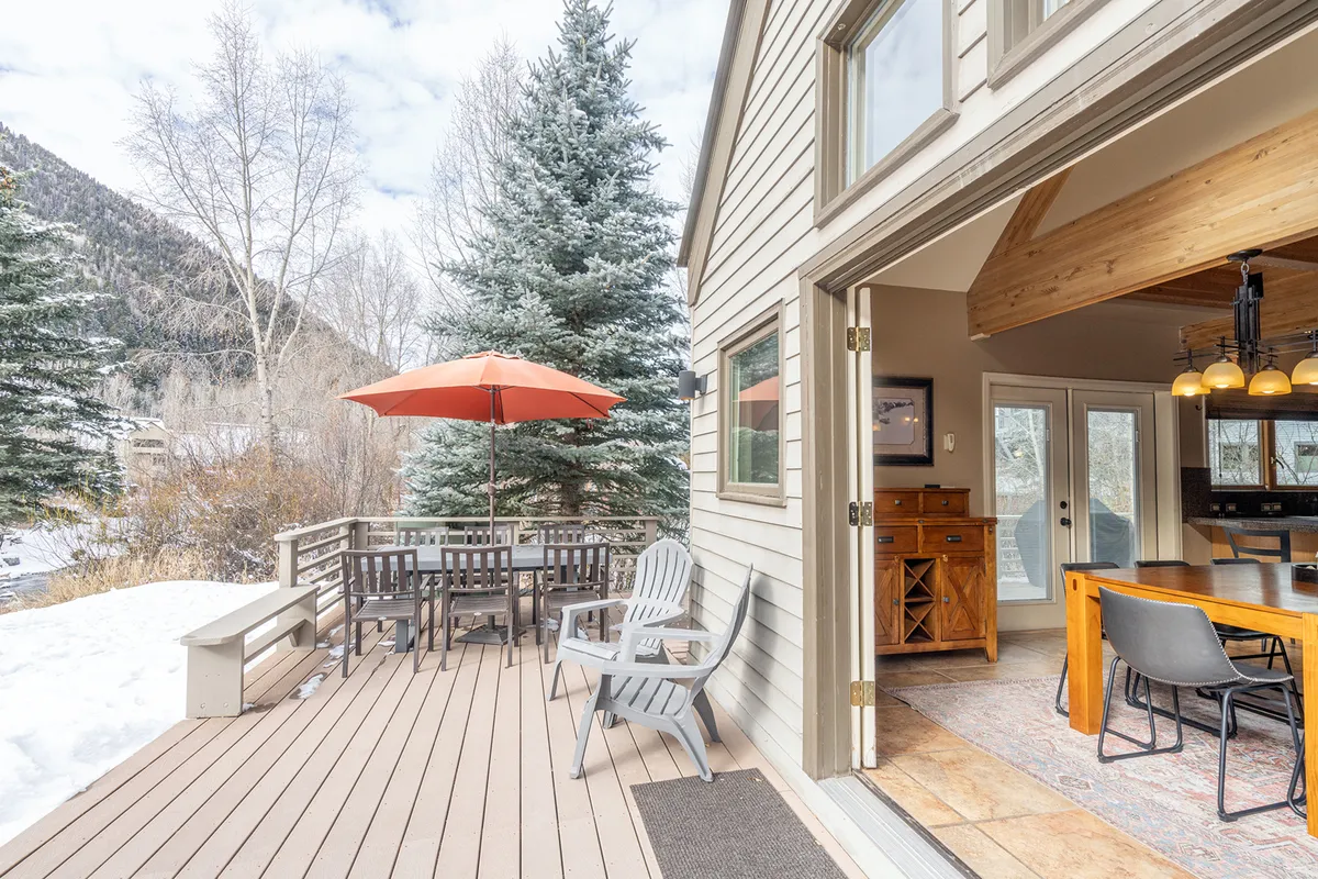 A spacious, well-furnished outdoor deck featuring a dining table shaded by an umbrella, several comfortable deck chairs, surrounded by lush trees, and opening to sweeping mountain views. The setting is perfect for al fresco dining, lounging, and social gatherings.