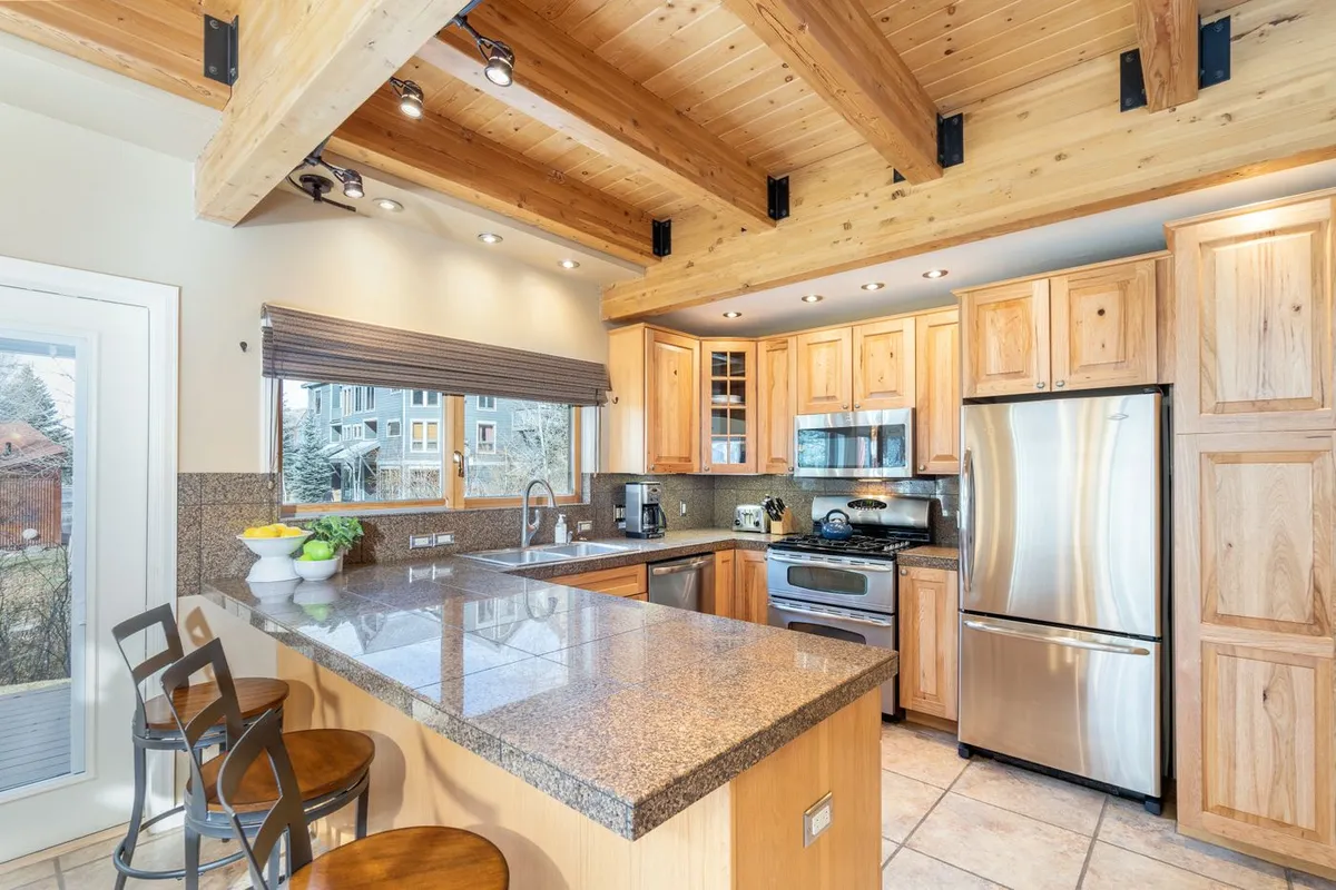 A bright, modern kitchen with a granite countertop serving as a spacious island, wooden beams overhead adding warmth, and sleek stainless steel appliances. The kitchen is open to the living space, with a window offering a pleasant view and natural light. There's ample space for meal prep and socializing, making it ideal for gatherings.