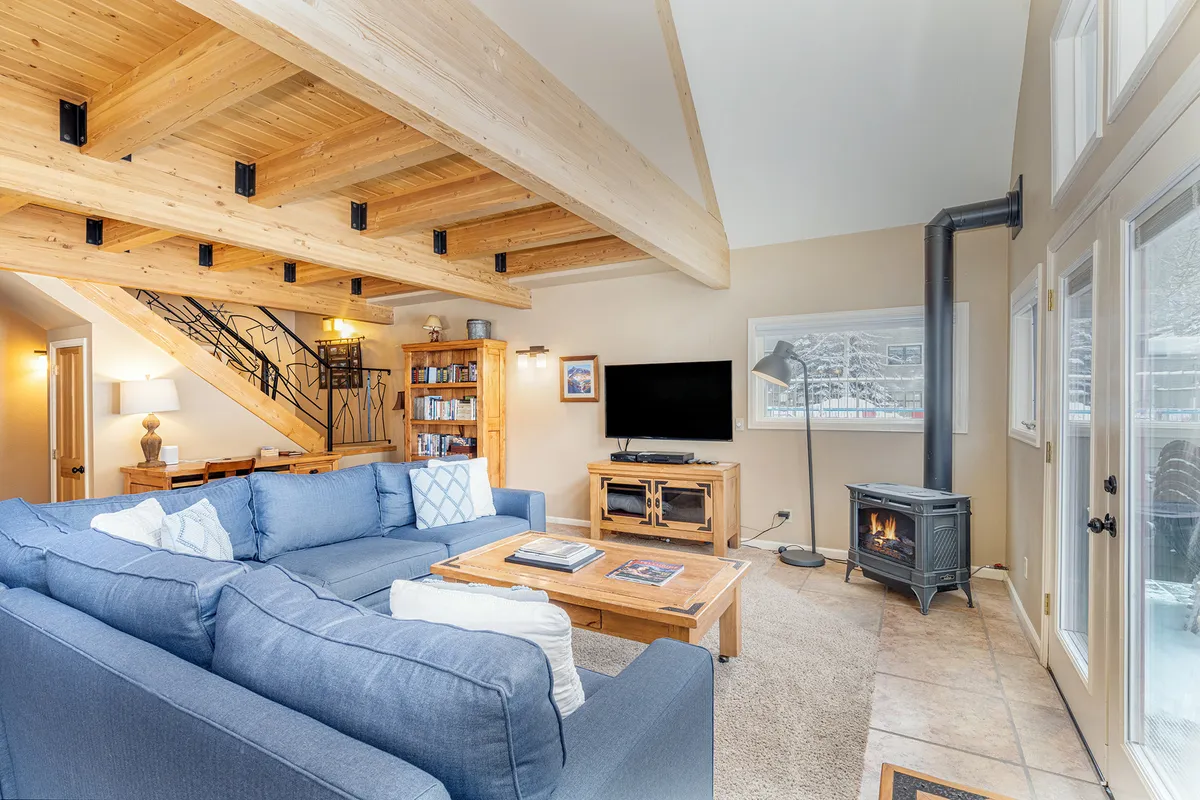 The living area showcases a large blue sectional sofa and a wooden coffee table set in front of expansive windows, inviting natural light and mountain views inside. The setting is ideal for movie nights, coffee mornings, or cozy conversations by the fire, emphasizing comfort and connection.