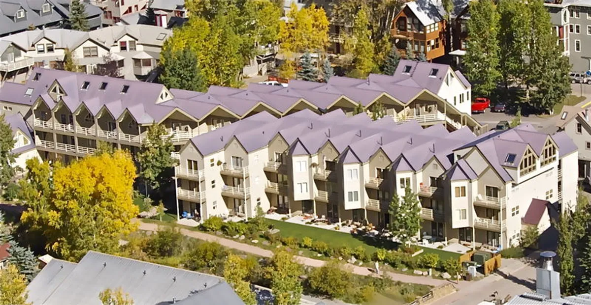 A row of attractive, multi-level townhouses is framed by lush green landscaping and vibrant autumn trees. Well-maintained pathways invite strolls and suggest easy access to community events and outdoor recreation.