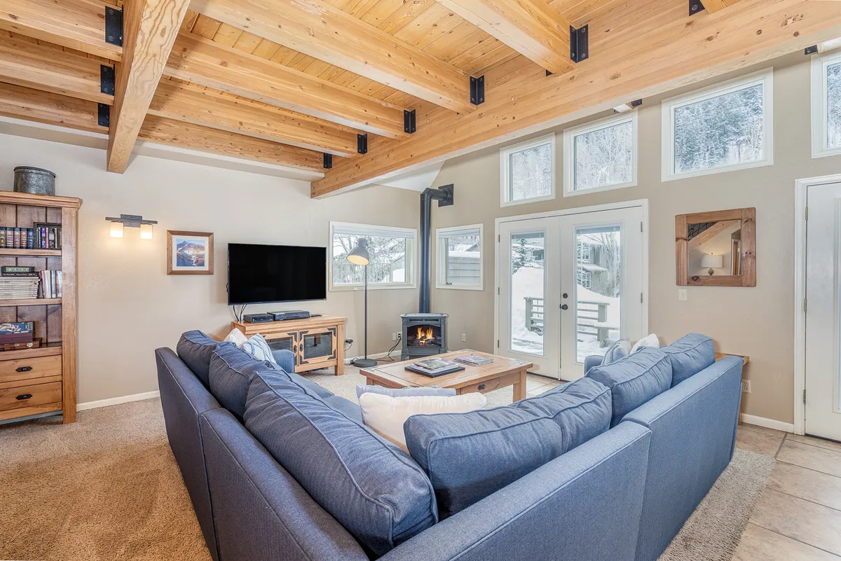 A spacious, welcoming living area featuring a large sectional sofa, exposed wooden beams, a cozy fireplace, and expansive windows. The space is open and connected to adjacent rooms, inviting guests to relax and gather after meals or during food prep.