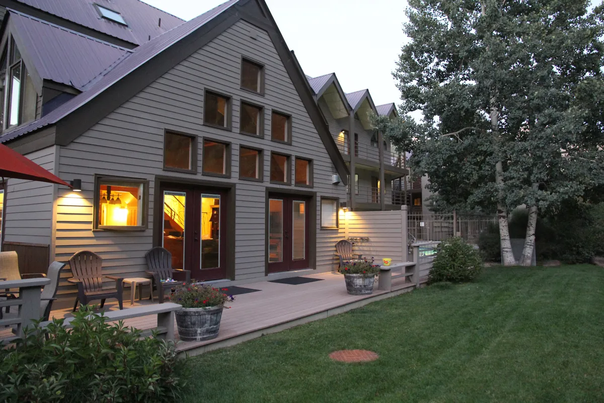 A generous wooden deck with comfortable seating overlooks a landscaped garden and features large windows connecting inside and out. The mood invites relaxation and socializing, with hints of evening barbecues or morning coffee surrounded by nature.