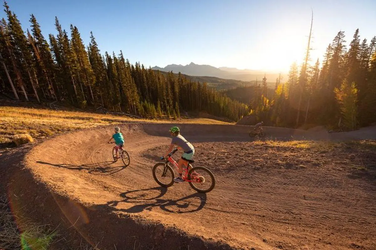 Mountain bikers ride along a winding trail through a dense forest at sunset, with distant mountains creating a dramatic backdrop. The scene is energetic and adventurous, capturing the spirit of active exploration in Telluride’s natural surroundings.