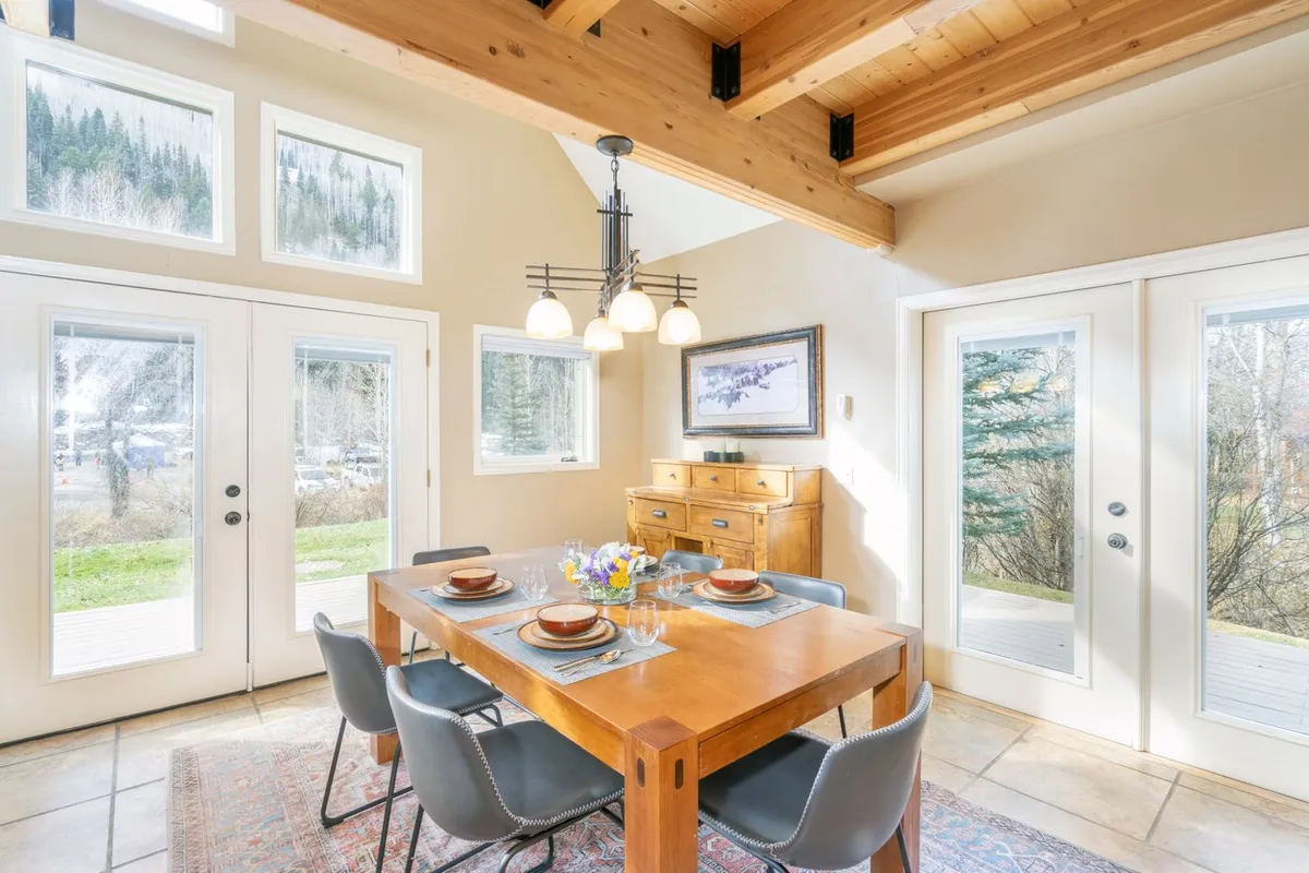 A bright dining area with a wooden table set for six, large windows letting in the sunlight, and rustic beams overhead. The sideboard and decorative painting enhance the inviting, contemporary mountain design. The image evokes shared meals, laughter, and togetherness, centered around the dining table.