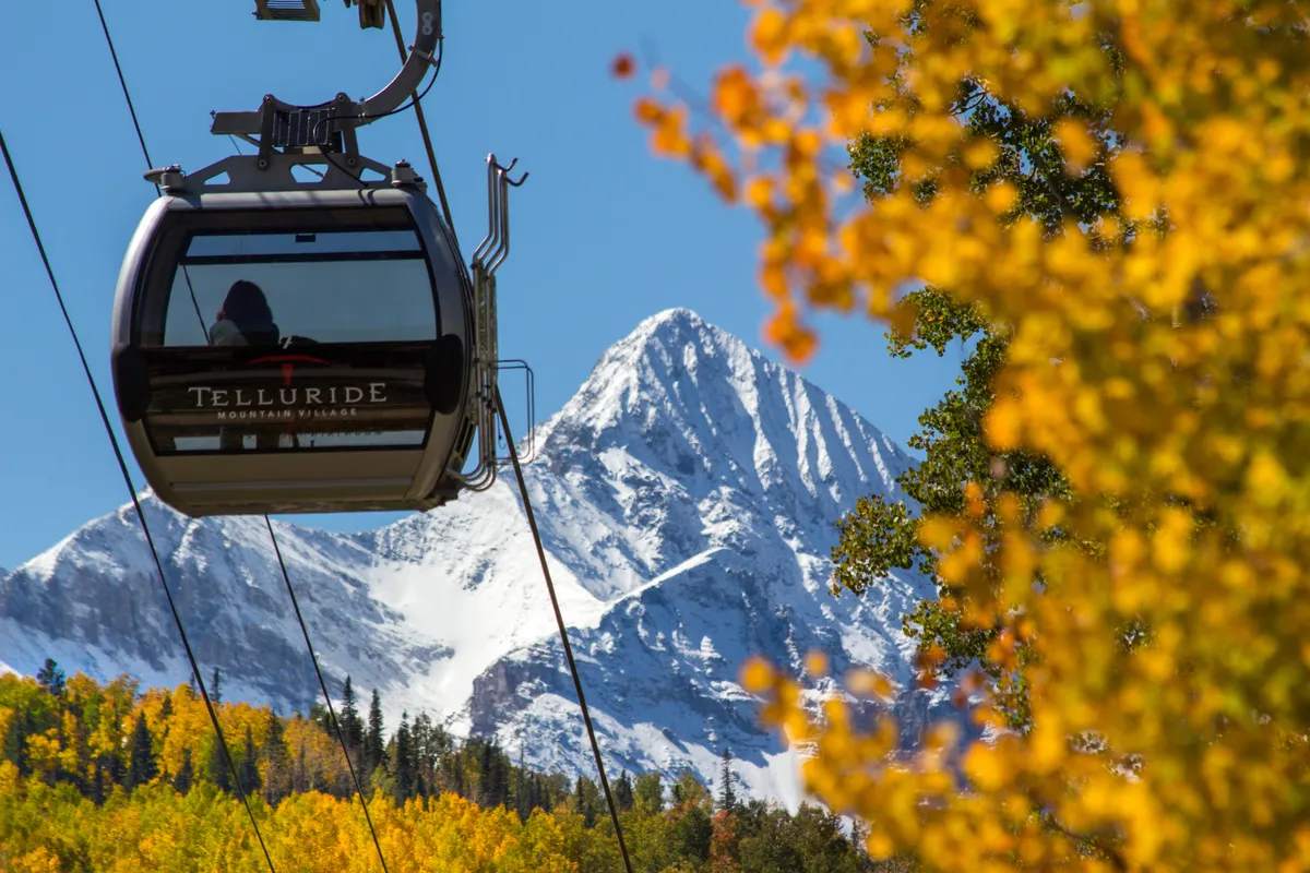 A gondola glides above the snow-capped mountains, with vibrant autumn foliage adding color to the scene. The view is expansive, highlighting Telluride’s dramatic landscape and easy access to both hiking and skiing via the gondola.