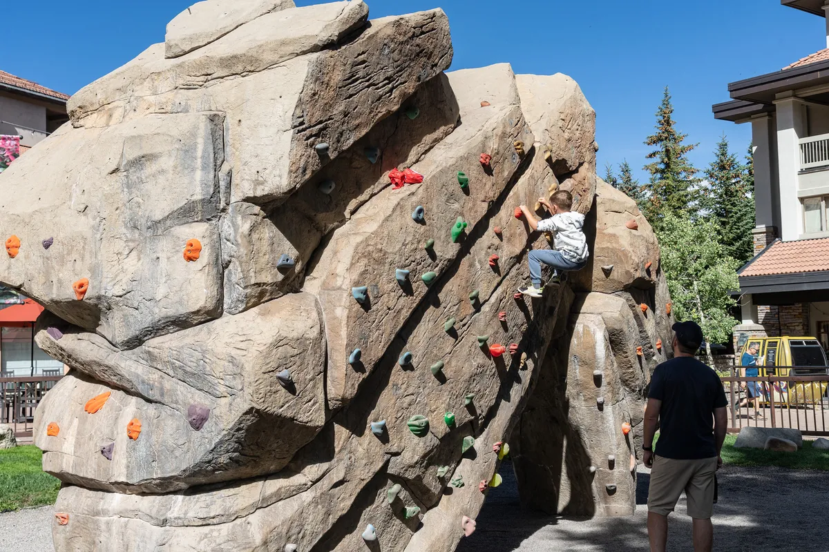 A child climbs a rock wall outdoors while an adult observes nearby, set against an active and energetic atmosphere. The climbing wall is surrounded by greenery, portraying safe, adventurous play and parental involvement—a great fit for highlighting kid-friendly activities.