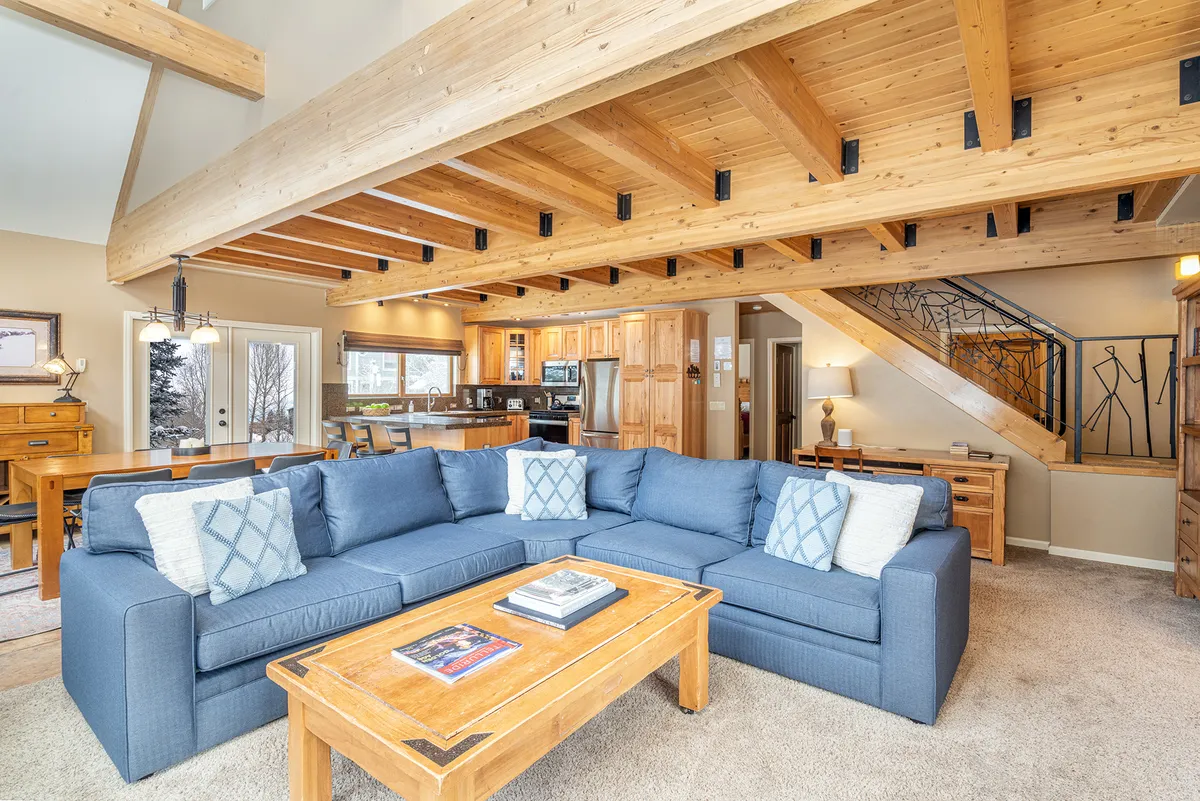 The image showcases a spacious, light-filled living area with a plush blue sectional sofa, a rustic wooden coffee table, and exposed wood beams on the ceiling. In the background, a modern kitchen is visible, creating an open-concept space perfect for gathering. The overall atmosphere is warm and inviting, blending mountain charm with modern comfort.