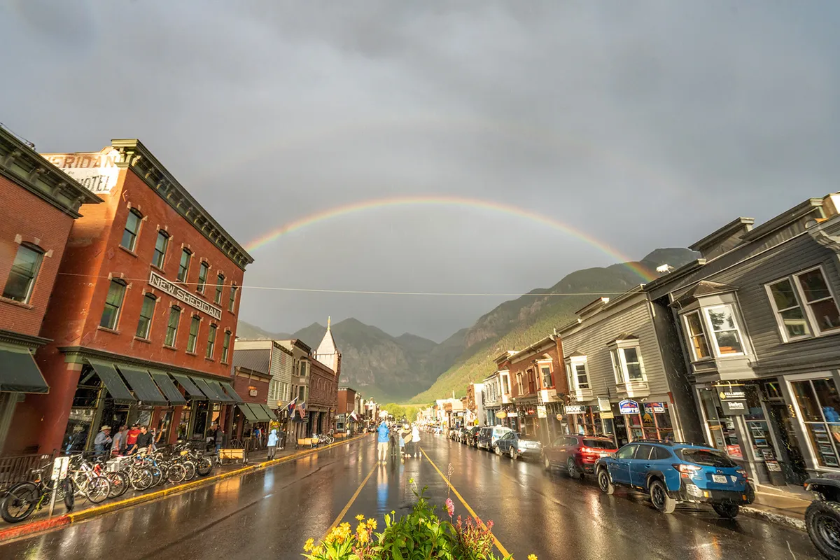 This image captures a vibrant downtown street scene with historic buildings, bicycles parked on the street, and a rainbow arching overhead. The atmosphere is lively and welcoming, suggesting a place full of unique local shops and cafes, ideal for exploration and spontaneous discovery.